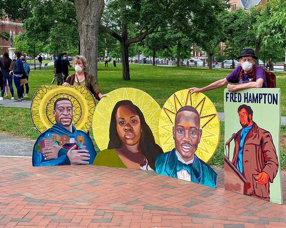 Protest art from David Fichter, right (muralist) pictured here with wife Debra Wise, (left), Artistic Director, Underground Railway Theater. The images portray unarmed Black victims of police murder: L–R, George Floyd; Breonna Taylor; Ahmaud Arbery—2020; and Fred Hampton, 1969. —photo by Marina Boni.